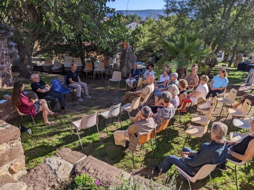 Dans un jardin, un groupe de 15 personnes est assis en demi cercle, ils font face à 3 personnes et une chaise vide