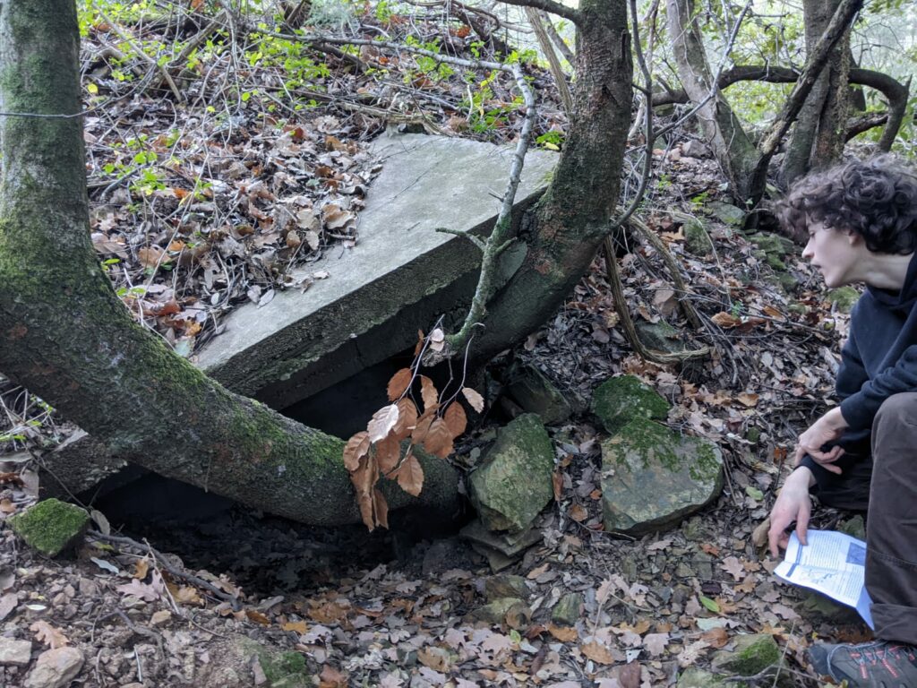 Sur la gauche, une dalle de béton posées de travers sur le sol d'une pente. Dalle en partie surélevée par les troncs de 2 arbres. Sur la droite un personnage une feuille à la main observe l'espace qui se trouve sous la dalle surélevée.