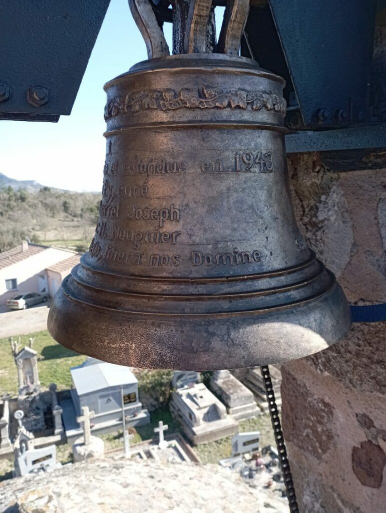 Gros plan sur la cloche de l'église. On peu lire une série d'inscription gravée sur la cloche. On distingue en arrière plan et au loin une maison et le cimetière qui se trouve à côté de l'église