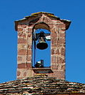 Vus sur le clocheton de l'église. Deux pillier avec un petit toit. Au milieu une arche et au sommet de l'arche est fixé la cloche de l'église. En bas de la photo on devine un bout de la toiture de l'église