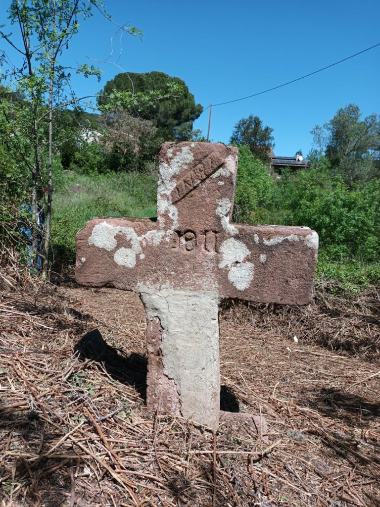 Une crois en pierre avec 2inscription: 1911 et au sommet INRI en travers. La croix a été consolidée avec un mortier à sa base. En arrière plan un terrain avec une importante végétation. Au loin des habitations et le ciel bleu