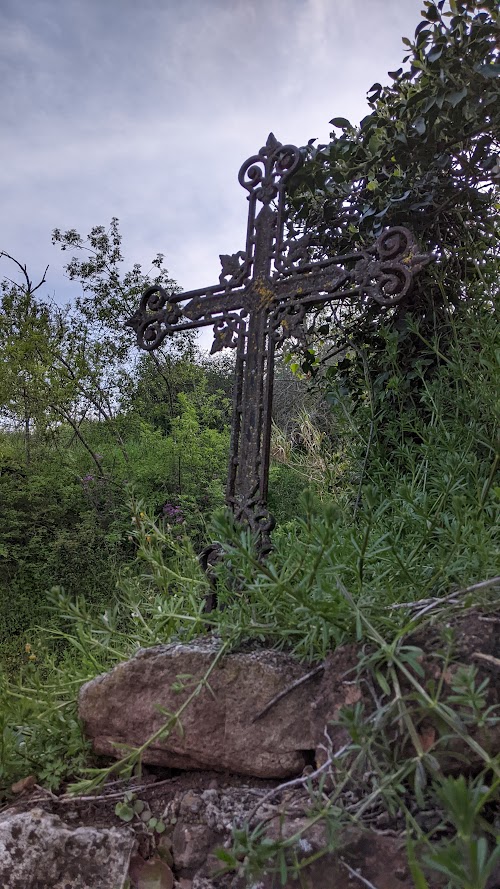 Une croix métallique rouillée avec des ornements. Les pointes de la croix sont décorées d'arabesques. La croix est planté sur le dessus d'un mur en pierre. En arrière plan beaucoup de végétation