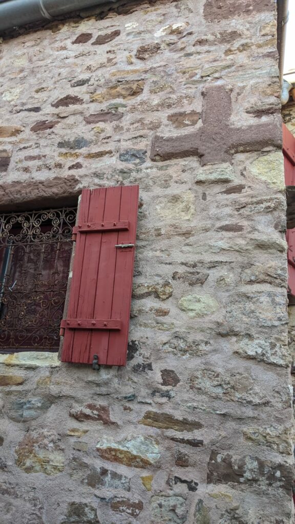 Vue sur la façade d'une maison en pierre de gré rouge. Les joints du mur ont été refait. Sur le côté une croix en gré rouge à été insérée dans le mur. Sur la gauche de la photo on voit une partie d'une fenêtre avec devant une grille en fer forgé et un volet en bois peint en bordeaux