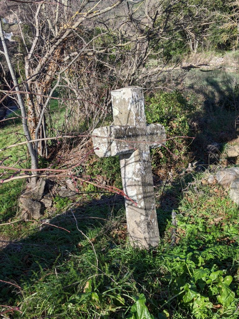 Une croix en pierre avec une inscription çàson sommet. La croix est penchée. Elle est planté dans la terre qui est recouverte de végétation et de fougères. En arrière plan de la photo, des jeunes pousse d'arbres