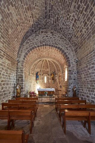 Vue sur l'intérieur de l'église. Belle voûte en pierre de gré rouge avec les joints refait à la chaux. Au fond le chœur de l'église avec des statues et l'autel recouvert d'une parure blanche. Au 1er plan les banc en bois avec une allée au milieu.