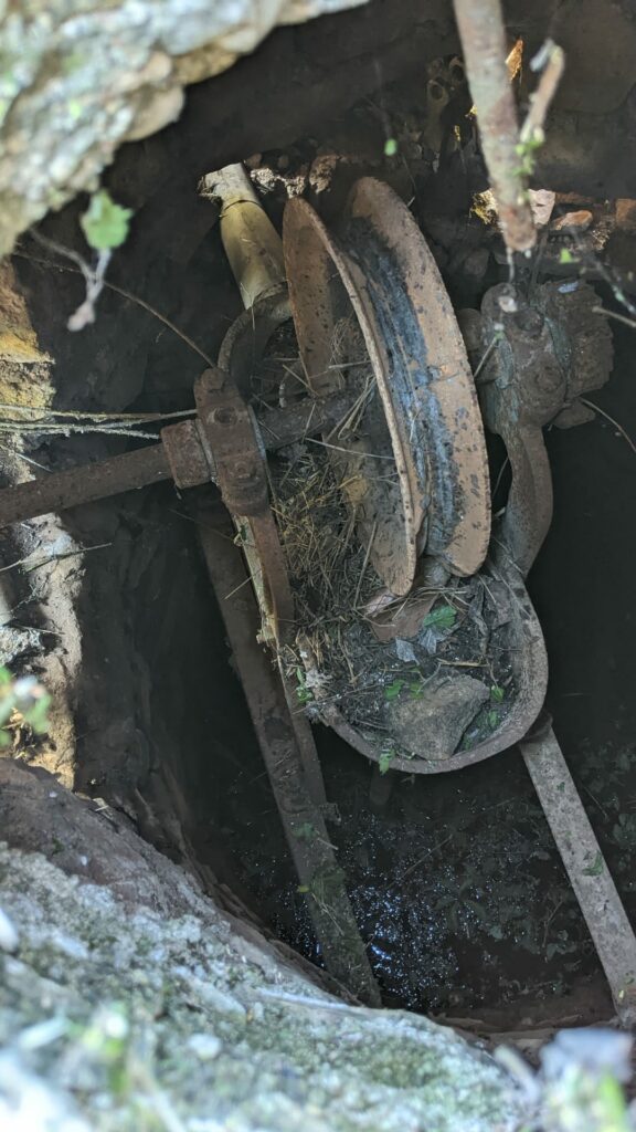 Photo prise depuis le trou situé sur la couverture de la construction en pierre protégeant un puits. Vue sur le mécanisme de fonctionnement du puits. Une roues, des pièces de métal, le tout est rouillé. Des cailloux et des herbes présents autour du mécanisme.