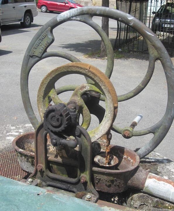 Photo du mécanisme d'une pompe à godet présenté sur une place goudronnée. Une grande roue-volant, devant une petite roue, des engrenages, une chaîne. En arrière plan des voitures garées.