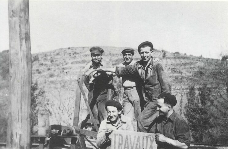 Photo en noir et blanc des années 50; Au fond une colline, on devine des terrasses. Au premier plan, 5 ouvriers. 3 sont montés sur une machine. 2 sont debout devant la machine ils tiennent un panneau sur lequel est inscrit le mot "travaux"