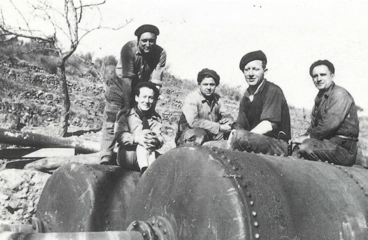 Photo noir et blanc des années 50. Au fond une colline avec des murs en pierres pour délimiter des terrasse. Au 1er plan 5 ouvriers assis sur 2 grosses citernes en métal. Les personnages ont tous un béret ou casquette sur la tête