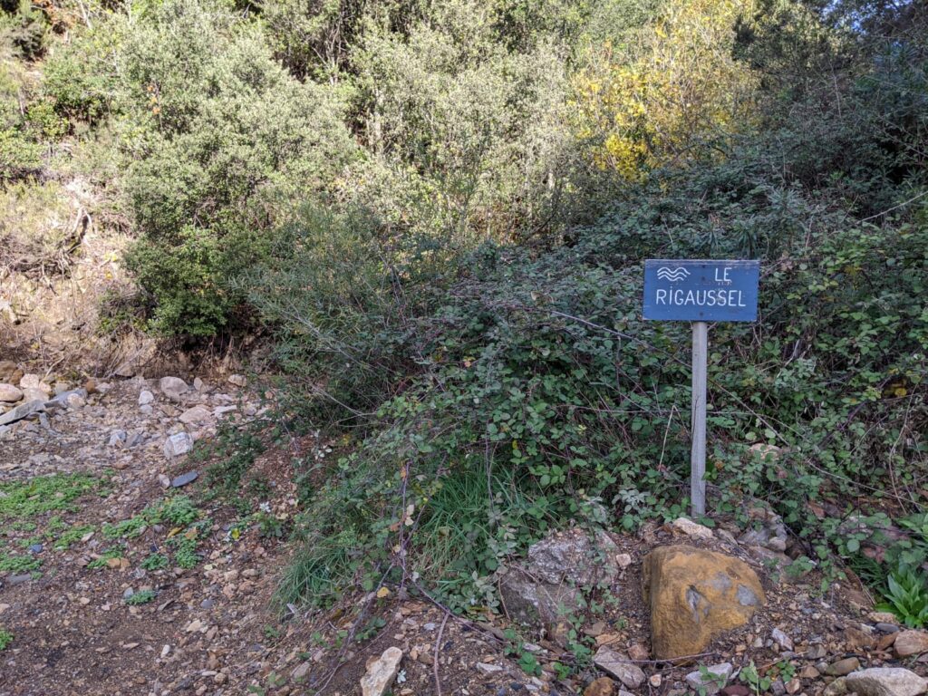 Au bord d'un chemin on devine le lit d'un ruisseau asséché qui sort de la végétation. Devant la végétation un panneau avec un pied métallique gris sur fond bleu sur lequel est écrit "Le Rigaussel"