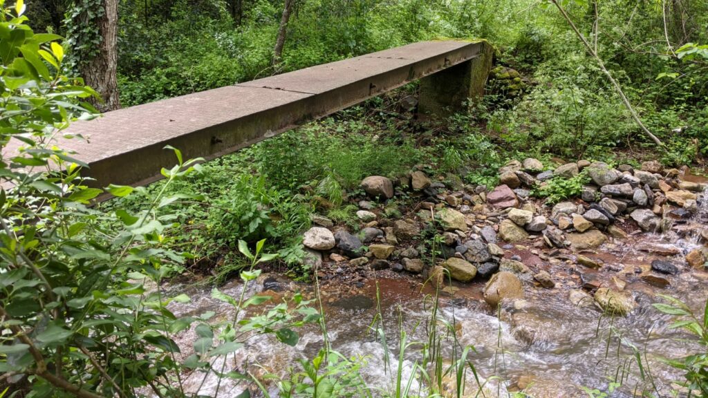 Vue sur un ruisseau avec de l'eau. Vue de biais sur une passerelle métallique. qui repose sur un pilier en béton ou en pierre. Sur les bords du ruisseau des pierres et sur les côtés du ruisseau une importante végétation