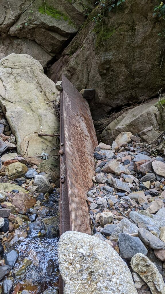 Dans le lit d'un ruisseau asséché, une passerelle métallique rouillée est bloquée entre des blocs de rochers et des tas de pierre