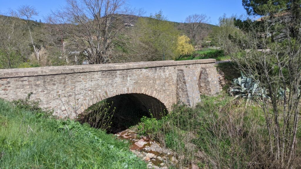 Un pont en pierre vue sur le côté. Le pont à une arche et 2 renforts en pierre sur le côté droit. Un filet d'eau coule sous le pont. De part et d'autre du ruisseau de l'herbe et des arbustes. Au fond on devine les montagnes des monts d'Orb