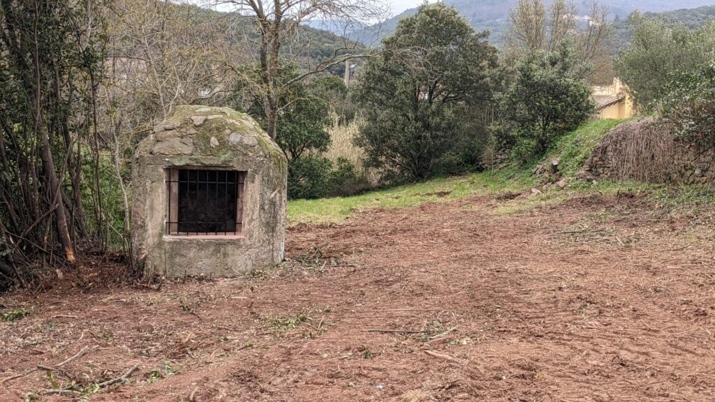 Vue sur la construction en pierre (environ 2 mètres de haut), dont le sommet est en forme de dôme, l'ensemble protège un puits. Sur le devant de la construction une ouverture d'environ 60 x 60 cm avec une grille de protection en fer. Le puits se trouve sur la gauche au milieu de la photo. devant la zone à été débroussaillé, il reste quelques branchages au sol. Derrière le puits, des arbres et quelques constructions sont visibles entre les arbres.