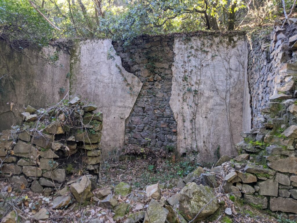 Vue de la ruine d'un bâtiment se trouvant à proximité de l'entrée de la galerie de la mine Verrière. Il n'y a plus de toit, le mur de façade est partiellement effondré, il y a des trace sur le mur du fond de l'emplacement d'une cheminée. Des feuilles et branchages sur le sol à l'intérieur du bâtiment.