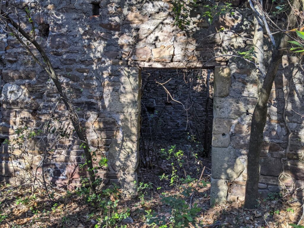 Vue de la ruine d'un bâtiment se trouvant à proximité de l'entrée de la galerie de la mine Verrière. Il n'y a plus de toit, sont visible, les murs, l'ouverture d'une porte. Des arbres ont poussé à l'extérieur et à l'intérieur du bâtiment.