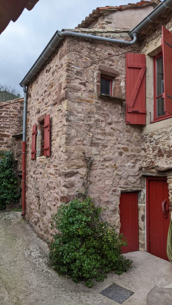 L'extension d'une maison en pierre avec 2 petite ouvertures (fenêtres) l'une dispose de volet l'autre non. En bas de cette construction une porte de petite taille. Volets et porte peinte en bordeaux. La construction est équipé de gouttière en zinc