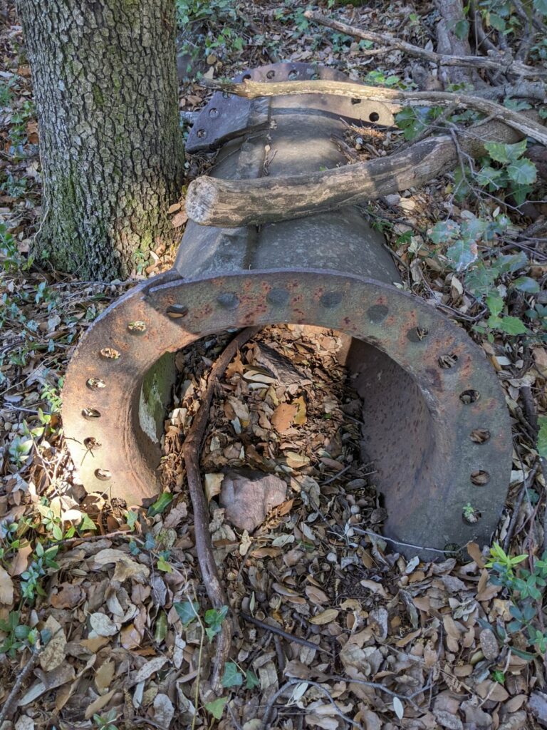 Un vieux tuyau rouillé d'environ 1 mètre posé sur le sol d'une foret au milieu des feuilles et branches près d'un tronc d'arbre