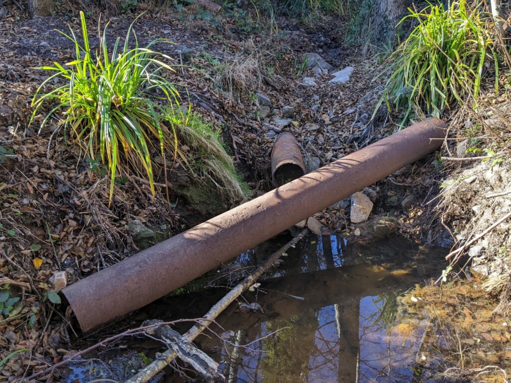 Le lit d'un petit ruisseau, en partie haute asséché, en partie basse une retenue d'eau. deux ancien tuyaux rouillée. Un dans le sens du ruisseau l'autre en traves. Sur chaque côtés du ruisseau un buisson d'herbe bien vert.