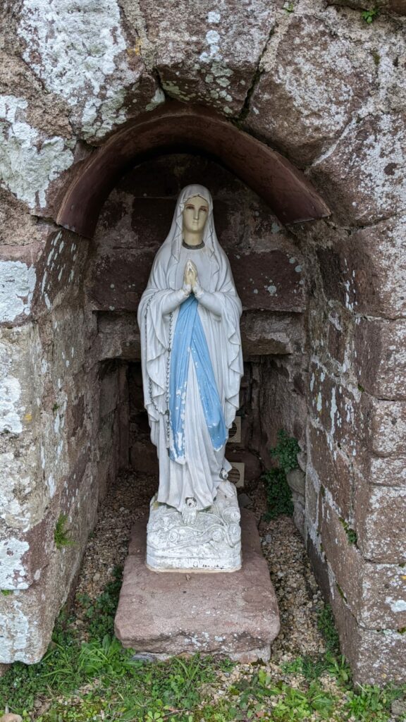 Une statue de la vierge Marie d'environ 50 cm de hauteur posée sur une pierre de grès rouge, autour de la pierre de l'herbe. La statue se trouve sous une arche de pierre de gré rouge.