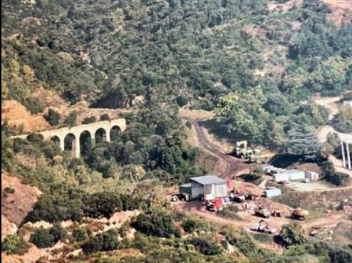 Une photo ancienne (années 80), un peu jaunie. Sur la gauche le viaduc d'Alzou. On voit arches. Sur la droite, une zone technique avec des bungalow, un hangar, des machines et des camions. Autour la végétation de la montagne