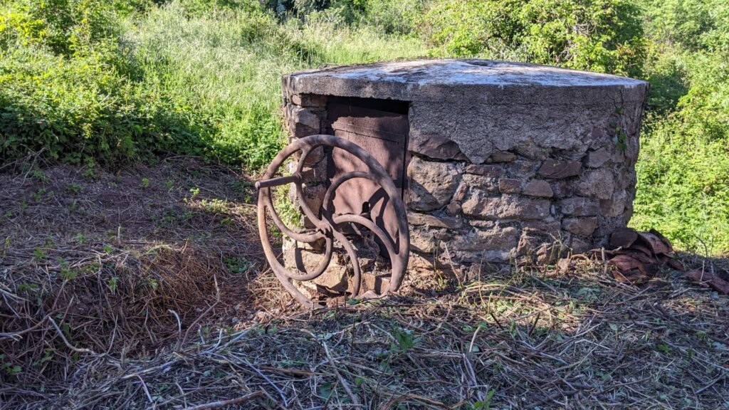 Vue sur la construction ronde, en pierre qui protège un puits. Sur le devant de la construction on peut voir la roue-volant du mécanisme de pompage. Derrière la construction de la végétation. Autour du puits des branchages au sol