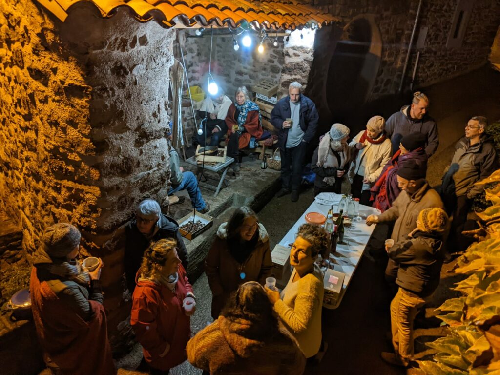 Un groupe de personne réunis dans une ruelle au milieu de laquelle est dressée une table. Il fait nuit. Les convives sont debout, certain un verre à la main. Il semble faire froid car les personnes sont chaudement habillées (certain avec des bonnets). La table à été dressée devant le fournil du four à pain de Frangouille. Une guirlande éclaire le fournil et la ruelle. Des personnes sont assises sur des bancs dans le fournil.